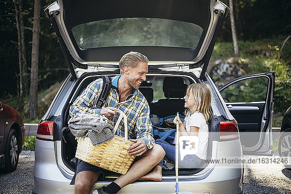 Lächelnder Vater mit Picknickkorb redet mit der Tochter  während er im Autokofferraum sitzt