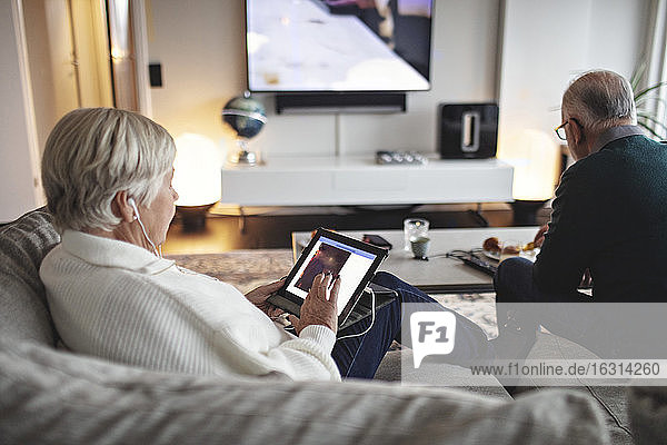 Ältere Frau benutzt digitales Tablett  während der männliche Partner auf dem Sofa im Wohnzimmer sitzt