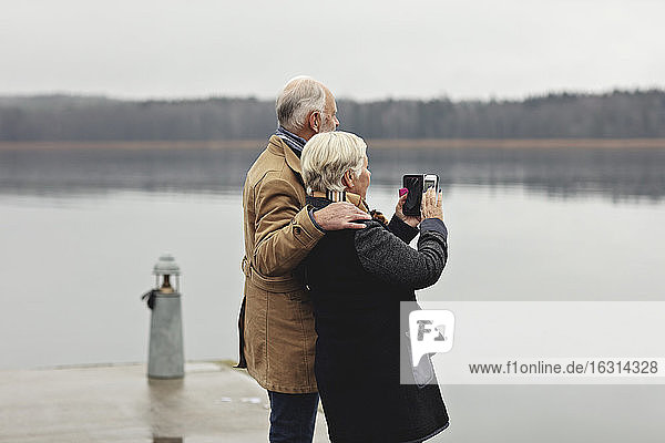 Senior couple photographing while standing by lake against clear sky