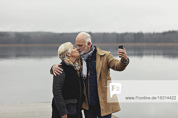 Senior couple kissing while taking selfie by lake against clear sky