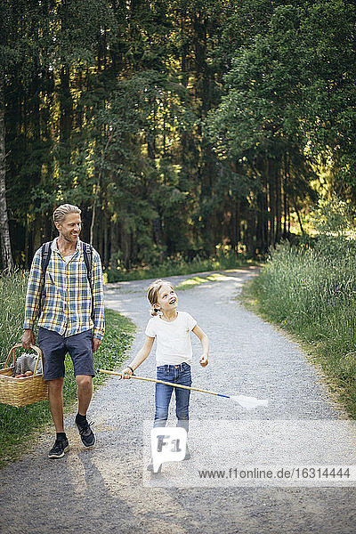 Smiling daughter talking to father holding picnic basket on road in forest