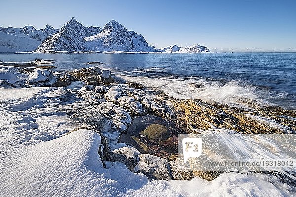 Verschneiter Felsstrand mit Gezeitentümpel  Küste mit blauem Meer  hinten winterliche Berge  Nordland  Lofoten  Norwegen  Europa