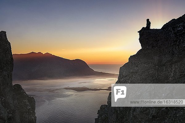 Silhouette einer Frau auf Felsvorsprung sitzend bei Sonnenuntergang  hinten Meer  Fjord und Berge mit Bodennebel  Gimsøy  Lofoten  Nordland  Norwegen  Europa