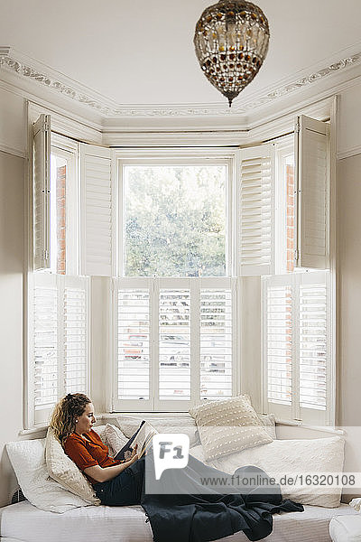 Young woman relaxing on living room sofa reading book in bay window