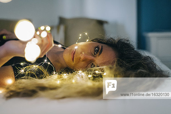 Porträt heitere junge Frau mit Lichterkette auf Bett