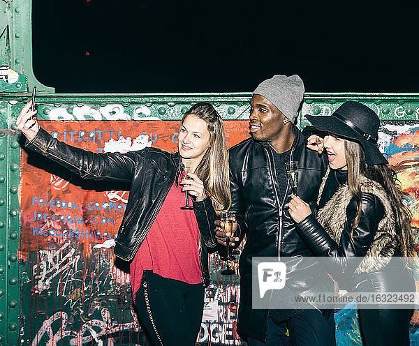 Freunde mit Sektgläsern machen nachts ein Selfie an einer Graffitiwand