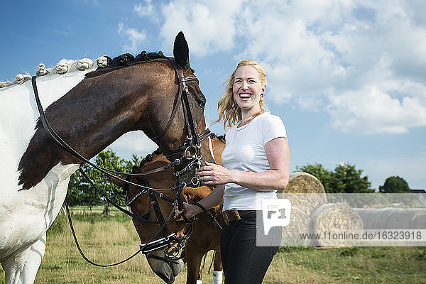 Deutschland  Wiesenau  lachende blonde Frau mit zwei Pferden auf einer Wiese