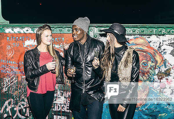 Friends holding champagne glasses standing at graffiti wall at night