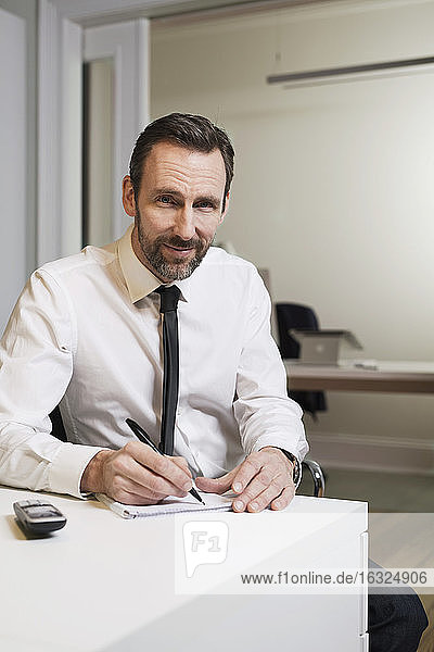 Portrait of confident businessman sitting at desk in office taking notes