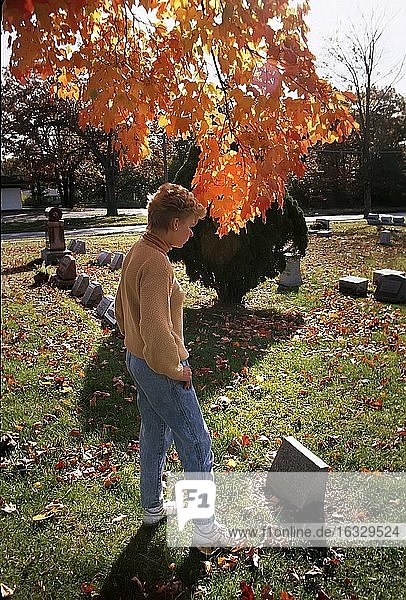 Female visits a grave site of a loved one in a cemetary with colorful tree in background.
