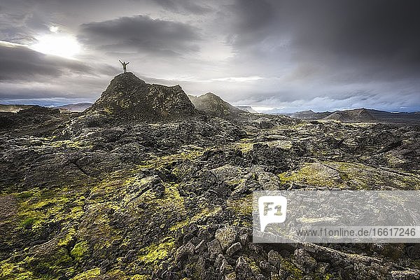Mann auf Hügel in einem schwarzen Lavafeld mit grünem Moos streckt Hände in den dramatischen Himmel  Leihrnjukur in der Krafla  Skútustaðir  Norðurland eystra  Island  Europa