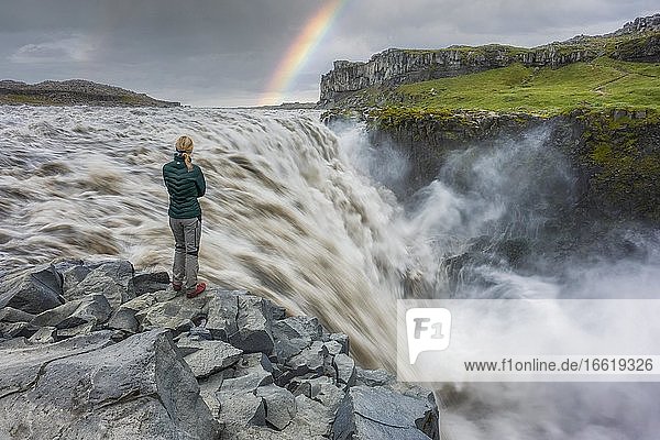 Blonde Frau mit grüner Daunenjacke steht am Dettifoss mit Regenbogen  Jökulsárgljúfur Nationalpark  Norðurþing  Norðurland eystra  Island  Europa