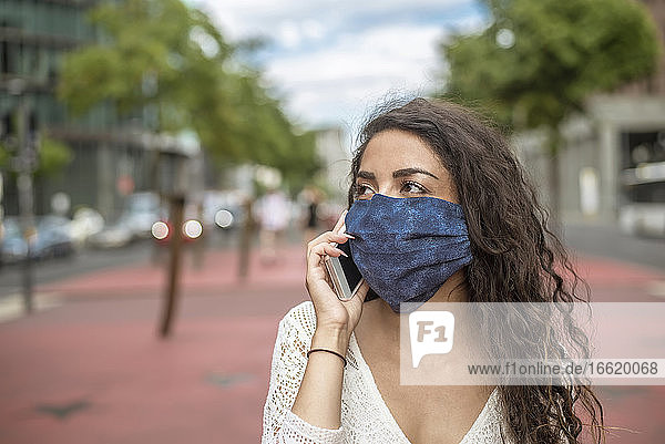 Nahaufnahme einer jungen Frau mit Maske  die mit einem Mobiltelefon in der Stadt spricht