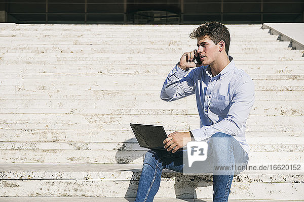 Gut aussehender junger Mann  der mit einem Laptop an einem sonnigen Tag sitzt und mit seinem Handy spricht