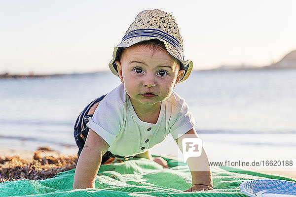 Nahaufnahme eines niedlichen kleinen Jungen mit Hut  der bei Sonnenuntergang auf einer Decke gegen das Meer krabbelt