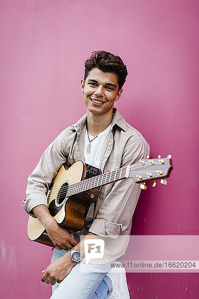 Glücklicher Mann hält Gitarre und lehnt sich an eine rosa Wand
