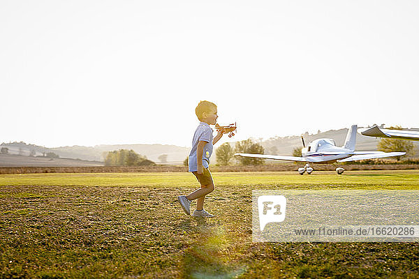 Niedlicher kleiner Junge  der mit einem Spielzeugflugzeug spielt  während er auf einem Flugplatz an einem sonnigen Tag steht