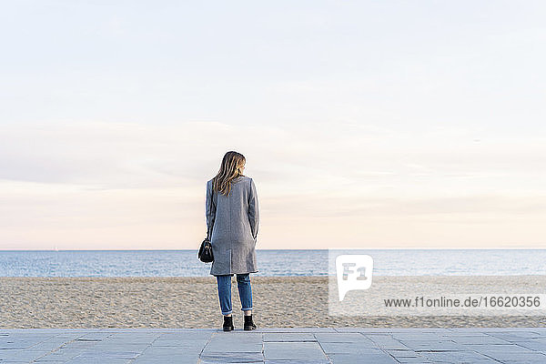 Junge Frau schaut auf das Meer  während sie auf der Promenade am Strand steht und den Sonnenuntergang genießt