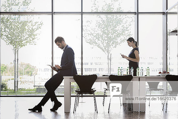 Geschäftsmann bei der Arbeit über einem digitalen Tablet  während eine Kollegin ihr Smartphone am Fenster im Büro benutzt