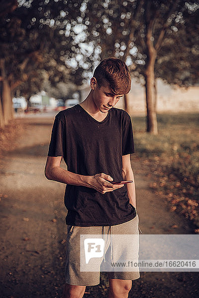 Teenage boy using mobile phone while standing on road during sunset