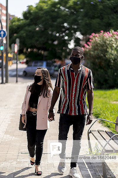 Junges Paar mit Gesichtsmasken hält sich beim Spaziergang in der Stadt während der COVID-19-Krise an den Händen