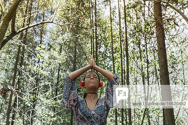 Junge Frau mit geschlossenen Augen  die über Kopfhörer Musik hört  während sie im Park gegen Bäume meditiert