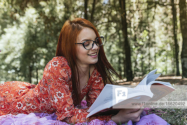 Lächelnde junge schöne rothaarige Frau  die ein Buch liest  während sie auf einer Picknickdecke mit Tagebuch im Park liegt