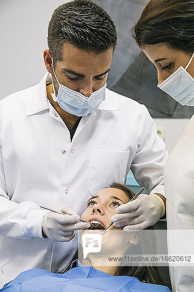 Männlicher Zahnarzt in Maske und Handschuhen untersucht die Zähne einer hübschen Patientin mit Hilfe einer weiblichen Assistentin in einer Klinik