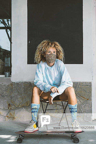 Frau mit Gesichtsmaske sitzt auf einem Stuhl mit Skateboard an der Wand