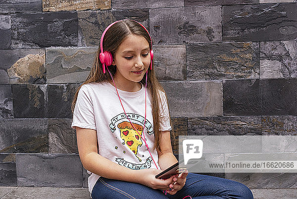 Nettes Mädchen mit Kopfhörern  das ein Mobiltelefon benutzt  während es zu Hause an der Wand sitzt