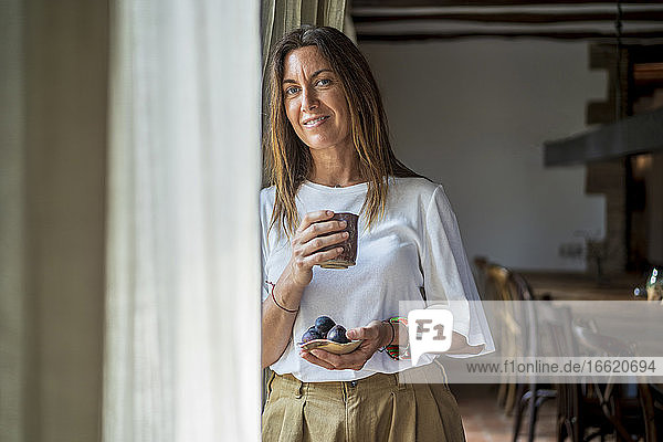 Lächelnde Frau mit Getränk und Obst in der Hand  die durch ein Fenster schaut