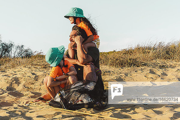 Vater und Töchter sitzen auf Sand mit Müllsack während eines sonnigen Tages