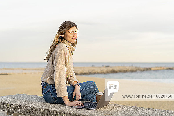 Nachdenkliche junge Frau  die mit ihrem Laptop auf einer Bank am Strand gegen den Himmel bei Sonnenuntergang sitzt und wegschaut