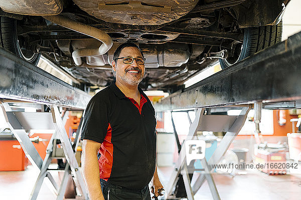 Lächelnder männlicher Mechaniker  der unter einem Auto in einer Autowerkstatt steht