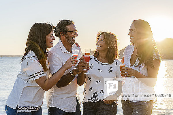 Eltern mit Töchtern stoßen mit Getränken an  während sie bei Sonnenuntergang am Meer stehen