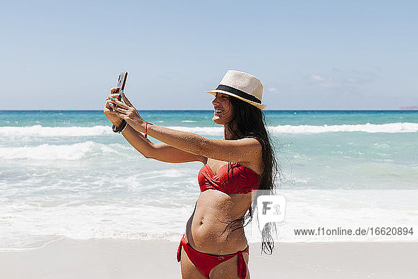 Frau im Bikini macht ein Selfie mit ihrem Smartphone  während sie am Meer steht