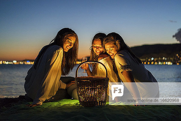 Lächelnde Schwestern betrachten einen beleuchteten Korb  während sie in der Abenddämmerung am Meer sitzen