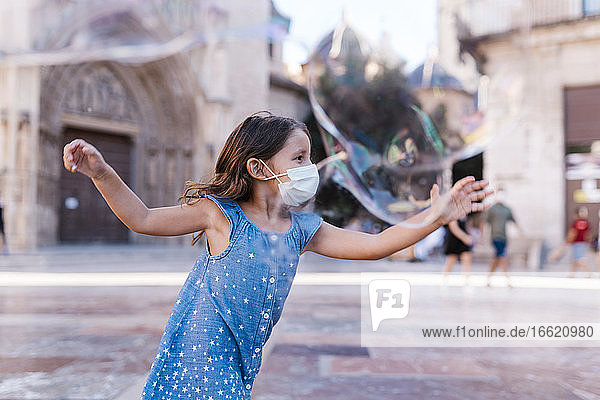 Carefree girl wearing mask running on street in city playing with soap bubble