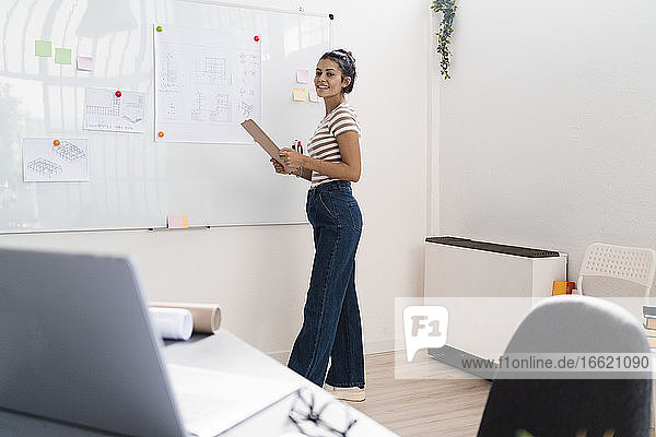 Lächelnde junge Architektin  die ein Klemmbrett hält  während sie an einem Whiteboard in einem kreativen Arbeitsumfeld steht
