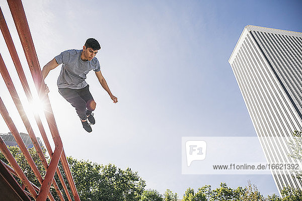 Junger Mann springt über Geländer Durchführung Parkour gegen klaren Himmel in der Stadt