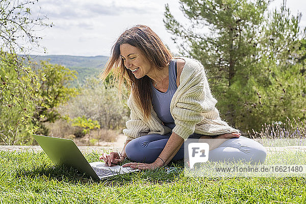 Lächelnde Frau  die einen Laptop benutzt  während sie im Hinterhof im Gras sitzt