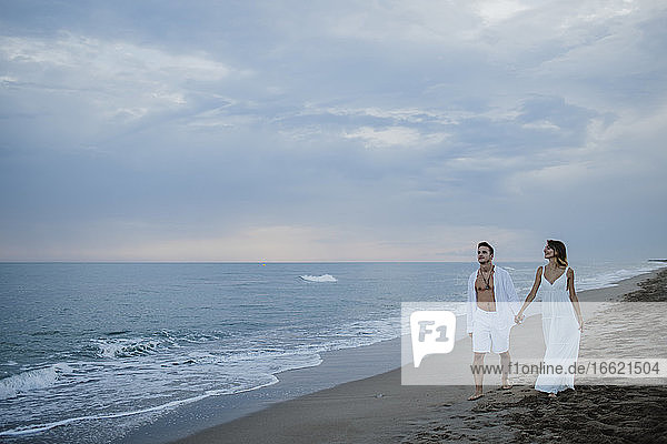 Junges Paar hält sich an der Hand  während es bei Sonnenuntergang am Strand spazieren geht