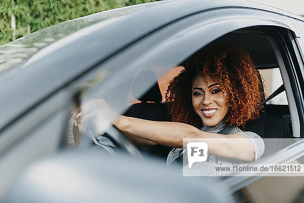 Lächelnde junge Frau mit Afrohaar am Steuer eines Autos  gesehen durch ein Fenster