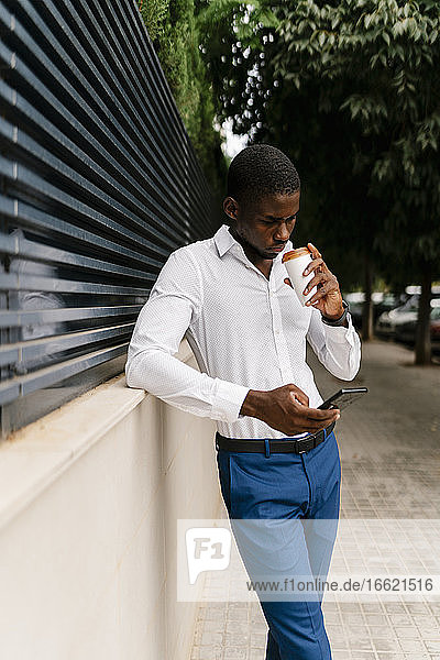 Geschäftsmann  der aus einer Tasse trinkt  während er sein Smartphone benutzt und sich an eine Wand lehnt