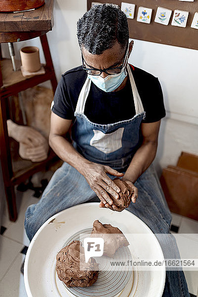 Männlicher Töpfer mit Maske  der Ton in der Hand hält und in einer Werkstatt sitzt