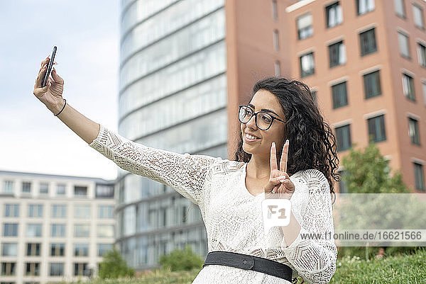 Lächelnde junge Frau zeigt das Friedenszeichen  während sie ein Selfie mit einem Smartphone in der Stadt macht