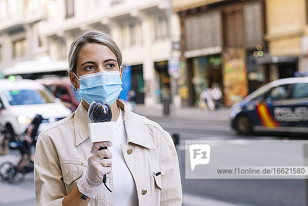Weibliche Journalistin mit Maske und Mikrofon schaut weg  während sie auf der Straße in der Stadt steht