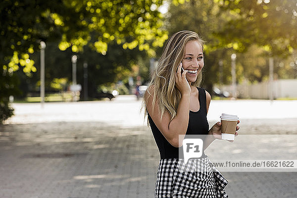Fröhliche junge Frau mit Kaffee in der Hand  die über ihr Smartphone spricht  während sie im Park steht