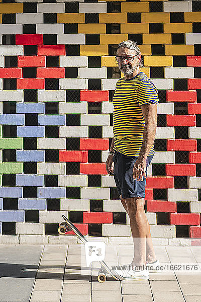 Mature man standing on skateboard against colored brick wall