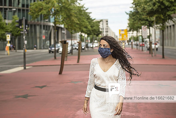 Junge Frau mit Maske schaut weg  während sie auf einem Fußweg in der Stadt spazieren geht